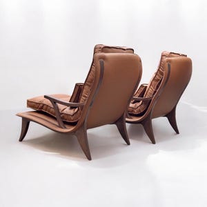 May include: Two brown leather lounge chairs with wooden frames. The chairs have a curved back and a wide seat. The chairs are facing away from the camera.