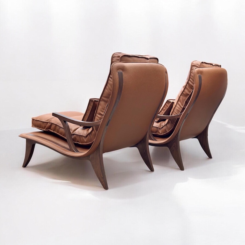 May include: Two brown leather lounge chairs with wooden frames. The chairs have a curved back and a wide seat. The chairs are facing away from the camera.