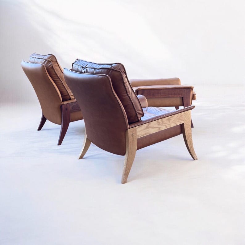 May include: Two brown leather armchairs with wooden frames and a matching ottoman. The chairs have a high back and are upholstered in a rich brown leather. The ottoman is also upholstered in brown leather and has a wooden frame.