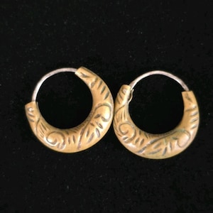 May include: A pair of hoop earrings with a brass-coloured finish and intricate etched designs. The earrings have a silver-coloured wire that goes through the ear. The background is a solid black.