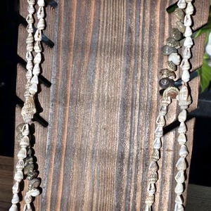May include: Two strands of seashell necklaces displayed on a wooden stand. The necklaces feature a variety of small, white seashells and some with gray and brown tones. The wooden stand has a dark brown finish with vertical wood grain.