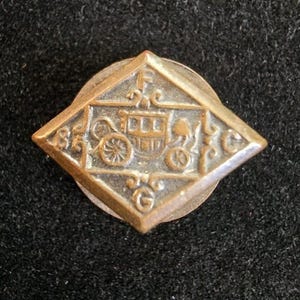 May include: A vintage brass pin with a diamond-shaped design. The pin features an embossed stagecoach and the number "85". The pin is set on a circular base and has an aged, antique appearance.