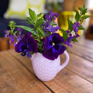 Puede incluir: Un ramo de flores moradas en una jarra de cerámica rosa pálido sobre una mesa de madera.