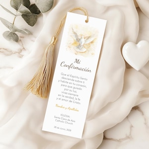 May include: A white bookmark with gold tassel, featuring a dove illustration and the text "Mi Confirmación". The bookmark includes religious text and the date "28 de Junio, 2028". A white heart-shaped decoration is visible in the background.