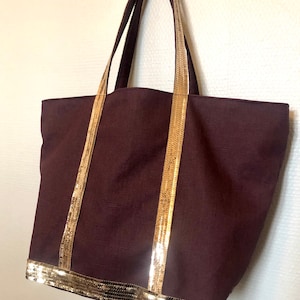 Puede incluir: Una bolsa de lona marr&oacute;n oscuro con ribete de lentejuelas doradas alrededor de la parte superior y las asas.