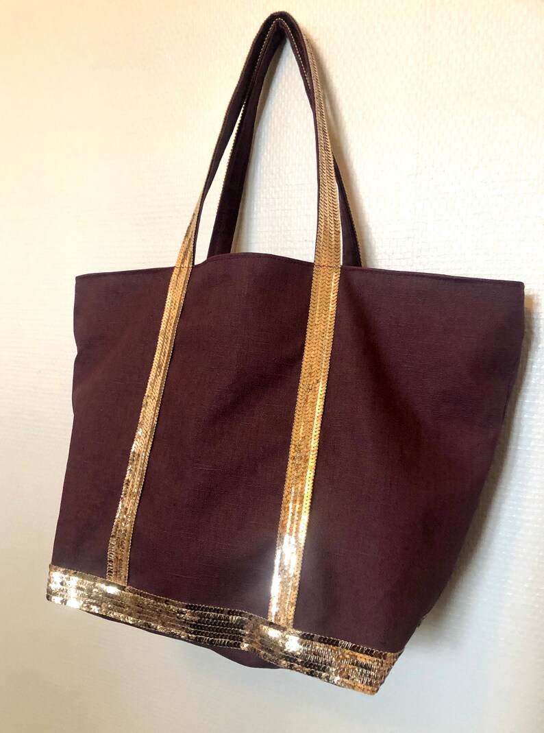 Puede incluir: Una bolsa de lona marr&oacute;n oscuro con ribete de lentejuelas doradas alrededor de la parte superior y las asas.