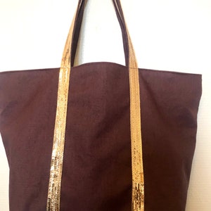 Puede incluir: Una bolsa de lona marr&oacute;n con ribete de lentejuelas doradas en las asas y la parte inferior.