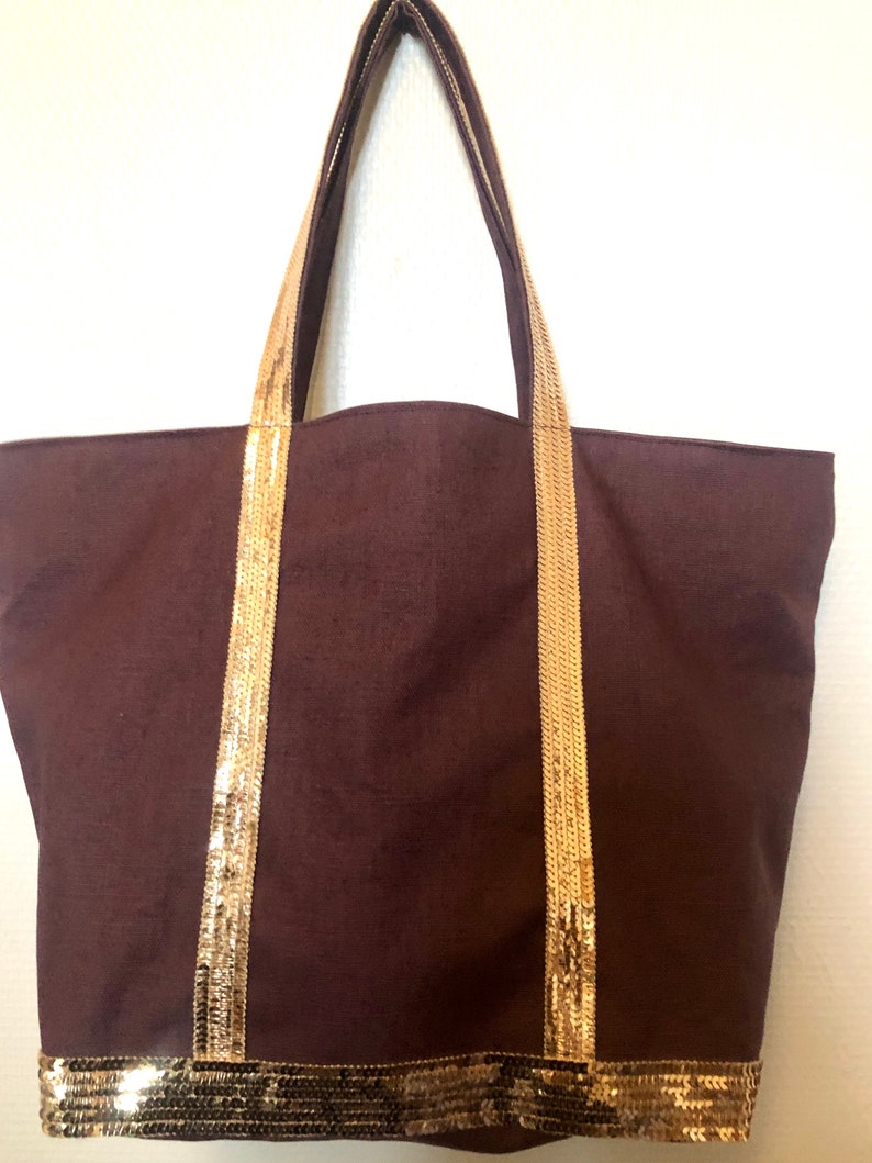 Puede incluir: Una bolsa de lona marr&oacute;n con ribete de lentejuelas doradas en las asas y la parte inferior.