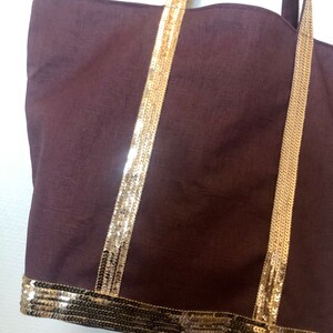 Puede incluir: Un bolso tote marr&oacute;n con ribetes de lentejuelas doradas en las asas y la parte inferior. El bolso est&aacute; hecho de una tela suave y texturizada.