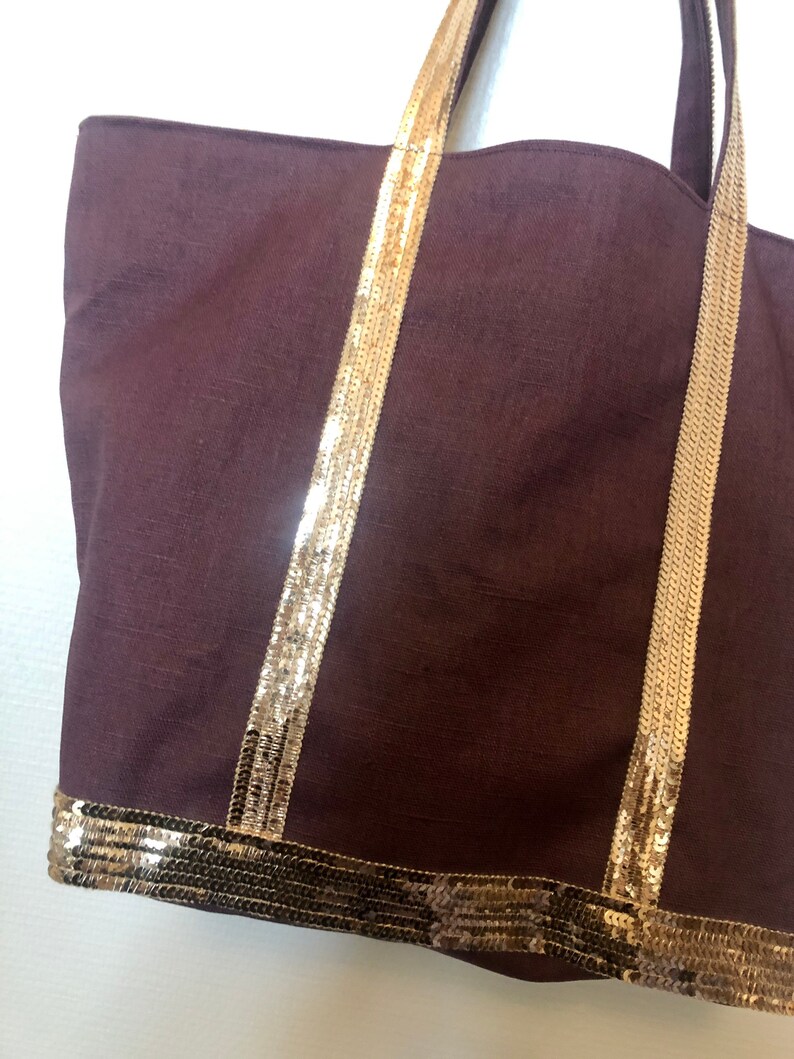 Puede incluir: Un bolso tote marr&oacute;n con ribetes de lentejuelas doradas en las asas y la parte inferior. El bolso est&aacute; hecho de una tela suave y texturizada.