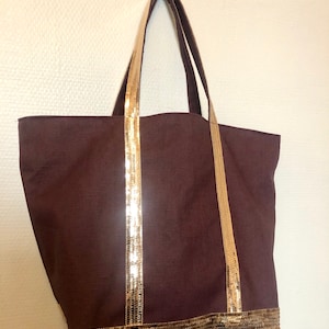 Puede incluir: Una bolsa de lona marr&oacute;n con ribete de lentejuelas doradas alrededor de la parte superior y las asas.