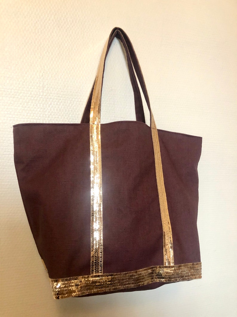 Puede incluir: Una bolsa de lona marr&oacute;n con ribete de lentejuelas doradas alrededor de la parte superior y las asas.