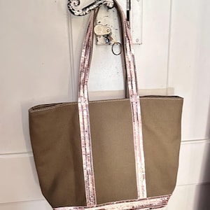 May include: A brown canvas tote bag with pink sequined straps and trim. The bag is hanging on a hook.