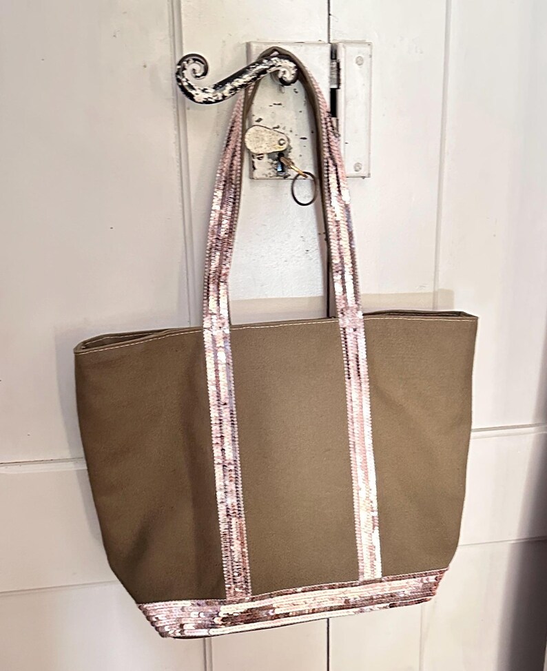 May include: A brown canvas tote bag with pink sequined straps and trim. The bag is hanging on a hook.