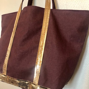 Puede incluir: Un bolso tote marr&oacute;n grande con ribetes dorados de lentejuelas en las asas y la parte inferior. El bolso est&aacute; hecho de una tela texturizada.