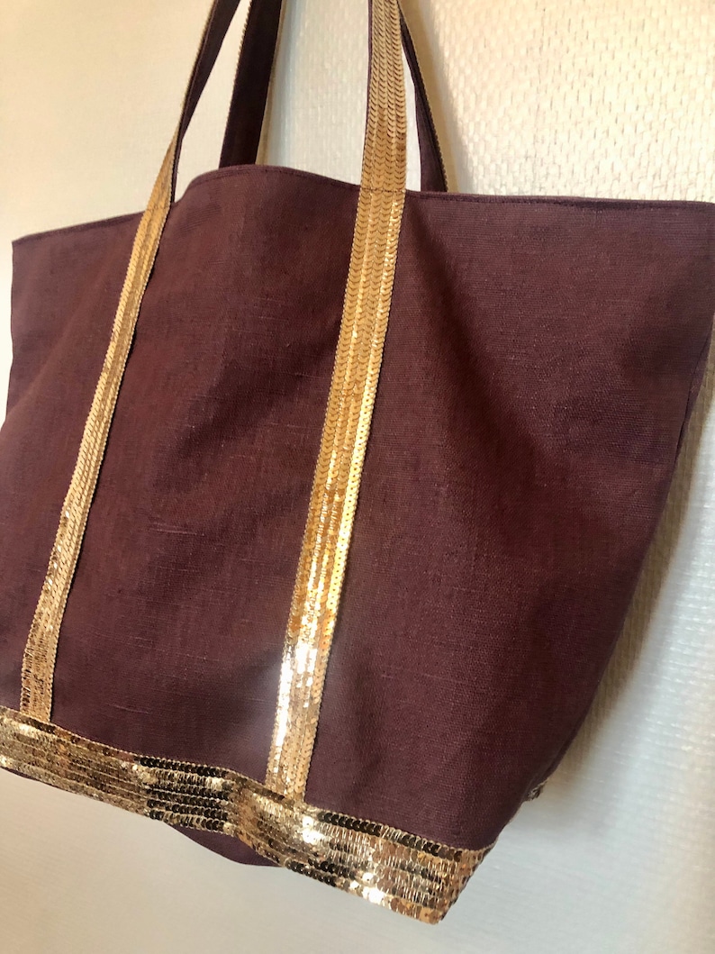 Puede incluir: Un bolso tote marr&oacute;n grande con ribetes dorados de lentejuelas en las asas y la parte inferior. El bolso est&aacute; hecho de una tela texturizada.