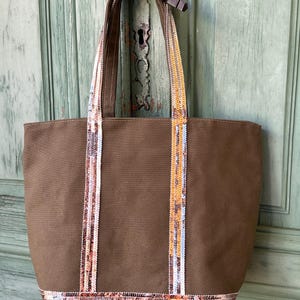 May include: A brown canvas tote bag with sequined straps and trim. The sequins are in shades of gold, pink, and white.