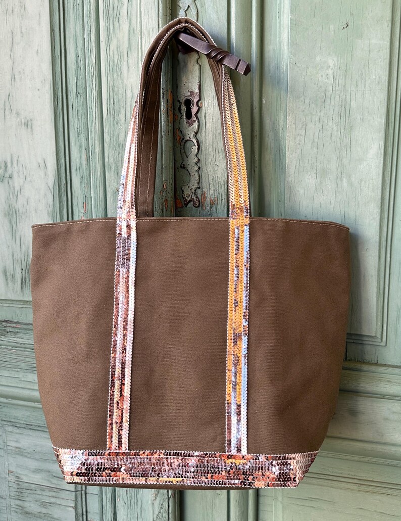 May include: A brown canvas tote bag with sequined straps and trim. The sequins are in shades of gold, pink, and white.