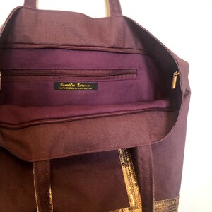 Puede incluir: Un bolso tote morado con ribete de lentejuelas doradas y un bolsillo con cremallera en el interior. El bolsillo tiene una etiqueta que dice "Camellia Forever" y una direcci&oacute;n de sitio web.