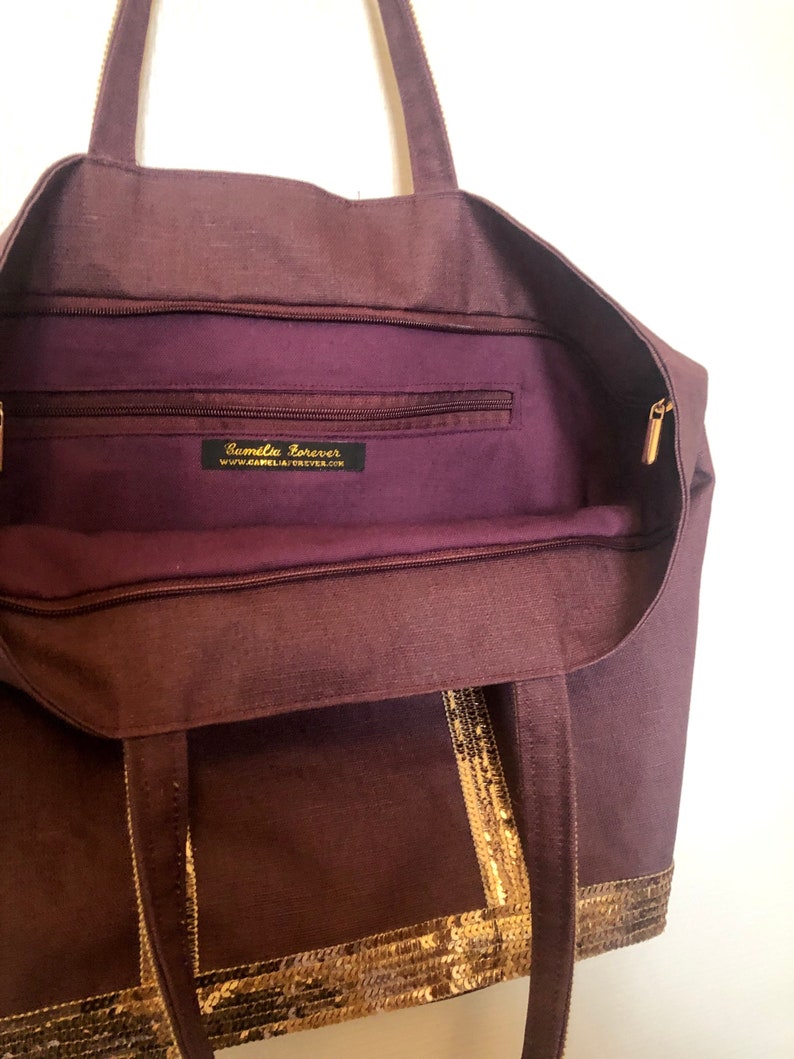 Puede incluir: Un bolso tote morado con ribete de lentejuelas doradas y un bolsillo con cremallera en el interior. El bolsillo tiene una etiqueta que dice "Camellia Forever" y una direcci&oacute;n de sitio web.