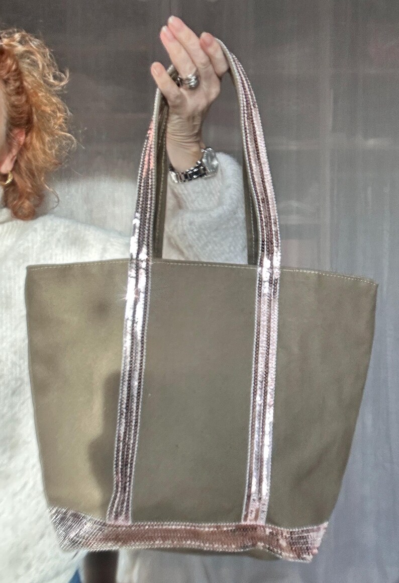 May include: A large, olive green canvas tote bag with pink and silver sequined straps and trim. The bag has a wide, flat bottom and two top handles.