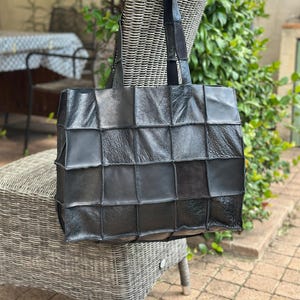 Op de afbeelding: Zwarte leren tote bag met een patchwork design. De tas heeft een rechthoekige vorm met een vierkant patroon en twee lange handvatten. De tas staat op een rieten stoel.