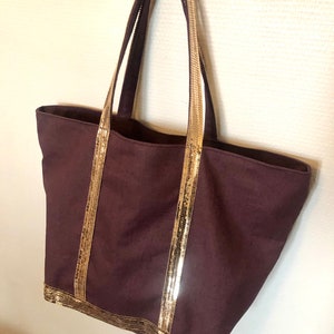 Puede incluir: Una bolsa de mano de color morado oscuro con ribete dorado de lentejuelas alrededor de la parte superior y las asas.