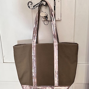 May include: A brown canvas tote bag with sequined straps and trim. The bag has two straps and a wide base.