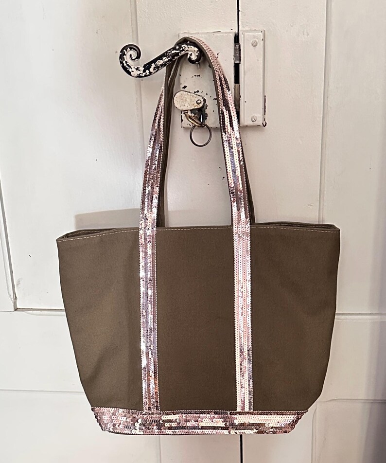 May include: A brown canvas tote bag with sequined straps and trim. The bag has two straps and a wide base.