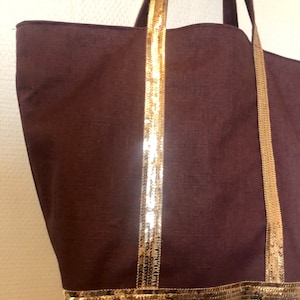 Puede incluir: Una bolsa de lona marr&oacute;n con ribete de lentejuelas doradas. La bolsa tiene dos asas con detalles de lentejuelas doradas.