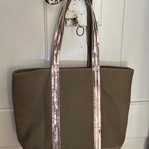 May include: A brown canvas tote bag with sequined straps and trim. The straps are a light pink and gold color.