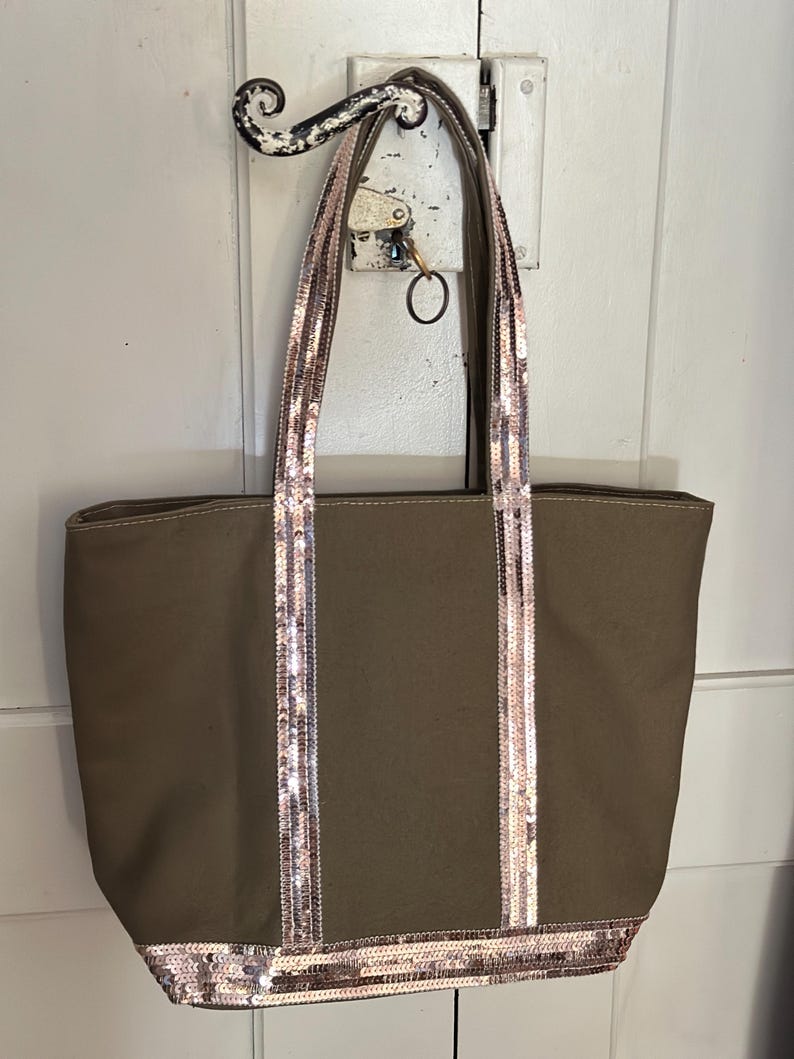 May include: A brown canvas tote bag with sequined straps and trim. The straps are a light pink and gold color.