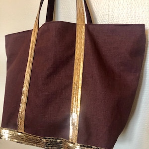 Puede incluir: Un bolso tote marr&oacute;n grande con ribetes dorados de lentejuelas en las asas y la parte inferior. El bolso est&aacute; hecho de un material similar al lino.