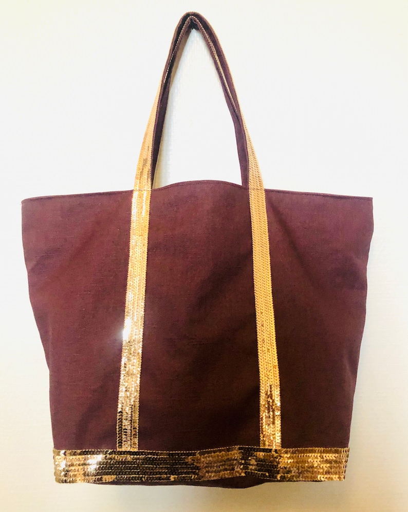 Puede incluir: Una bolsa de lona marr&oacute;n con ribete de lentejuelas doradas. La bolsa tiene dos asas largas y una base ancha.