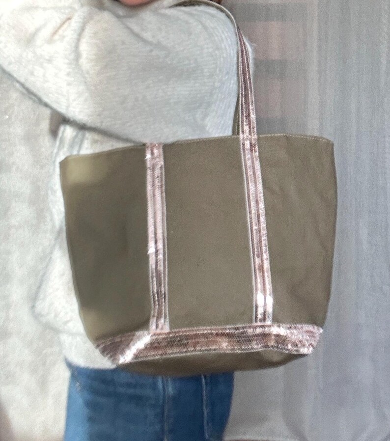May include: A large, olive green canvas tote bag with pink and white sequined trim. The bag has two long handles.