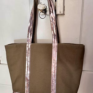 May include: A brown canvas tote bag with pink and silver sequined straps and trim. The bag is hanging on a hook.