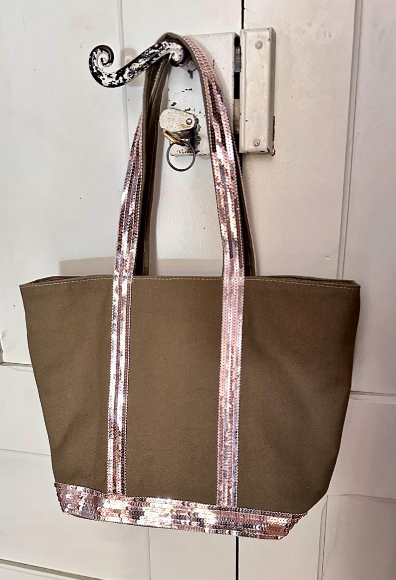 May include: A brown canvas tote bag with pink and silver sequined straps and trim. The bag is hanging on a hook.