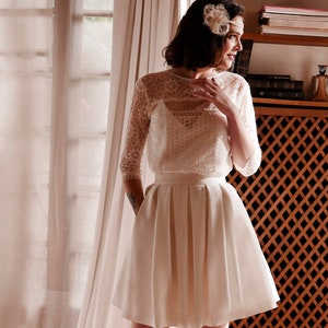 Op de afbeelding: Een vrouw draagt een wit kanten topje en een witte satijnen rok. Ze staat voor een raam met witte gordijnen en een boekenkast achter haar.