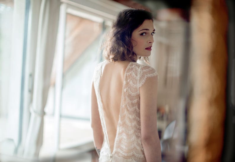 May include: A woman wearing a white lace wedding dress with a low back and a plunging neckline.