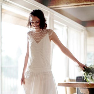 May include: A woman wearing a white lace dress with a scalloped neckline. She is smiling and holding a bouquet of white flowers with green leaves.