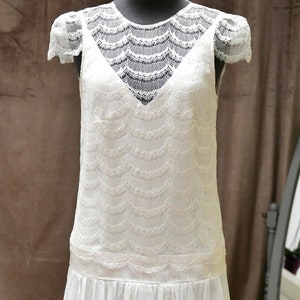 May include: A white lace dress with a scalloped lace pattern and a v-neckline. The dress has a fitted bodice and a flowing skirt.