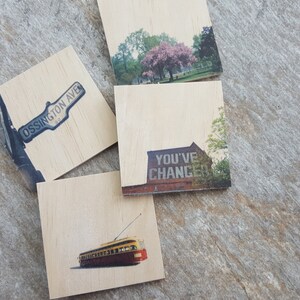 May include: Four wooden coasters with different images printed on them. The coasters feature a street sign that says "Ossington Ave", a building with the words "You've Changed" painted on it, a cherry blossom tree in a park, and a vintage streetcar.