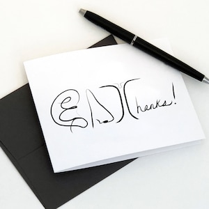 May include: A white card with a black line drawing of a face and the word "thanks!" written in black ink. The card is sitting on a black envelope.