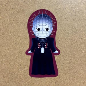 May include: A red and black sticker featuring a cartoon character with a spiky, wire-mesh head and a black dress. The character has a white face with large, black eyes.