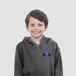 Pu&ograve; includere: Un giovane che indossa una felpa con cappuccio grigia scura con un cappello con orecchie di Topolino ricamato in blu e bianco sul davanti.
