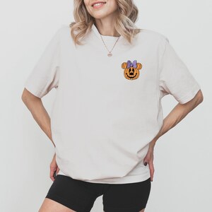 May include: A white t-shirt with a pumpkin-shaped Mickey Mouse head with a purple bow on it.