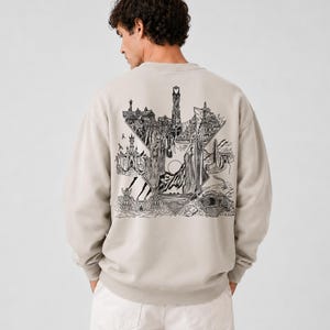 May include: Beige sweatshirt with a black and white graphic design on the back. The graphic depicts a detailed landscape with buildings, mountains, and figures. The sweatshirt is a neutral color and has a crew neck.