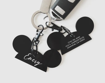 May include: Black Mickey Mouse-shaped keychains with a white inscription. One keychain says "Enery" and the other has a phone number, address, and city.