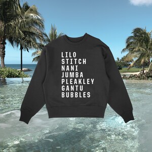 K&ouml;nnte beinhalten: Schwarzes Sweatshirt mit Rundhalsausschnitt und wei&szlig;em Text mit den Namen: Lilo, Stitch, Nani, Jumba, Pleakley, Gantu und Bubbles. Das Sweatshirt wird vor einem tropischen Strandhintergrund mit Palmen und Meer gezeigt.