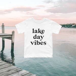 May include: White t-shirt with black text that reads "lake day vibes" against a backdrop of a lake and a wooden dock.